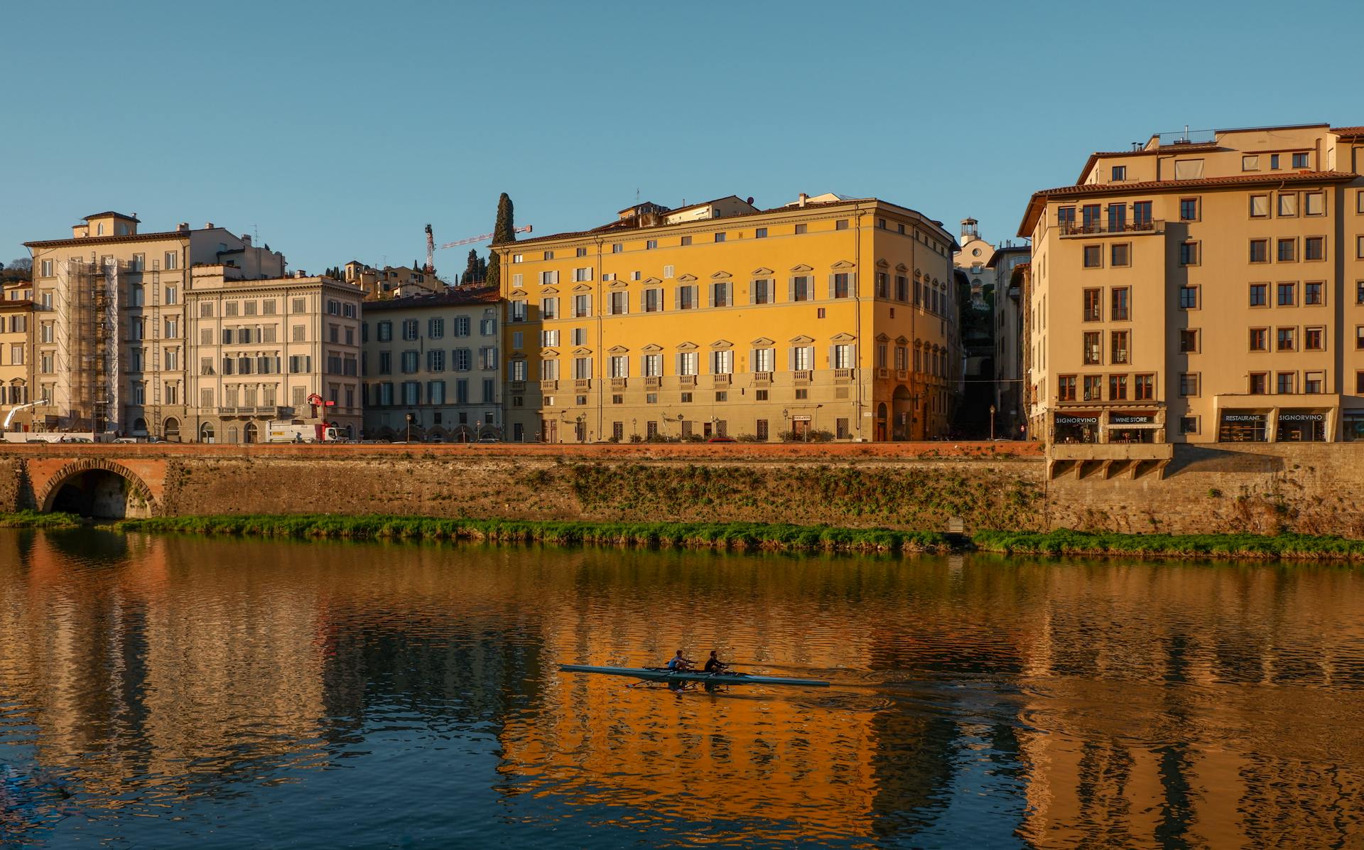 Rowers canoe along a river with historic buildings reflecting under a clear blue sky.
