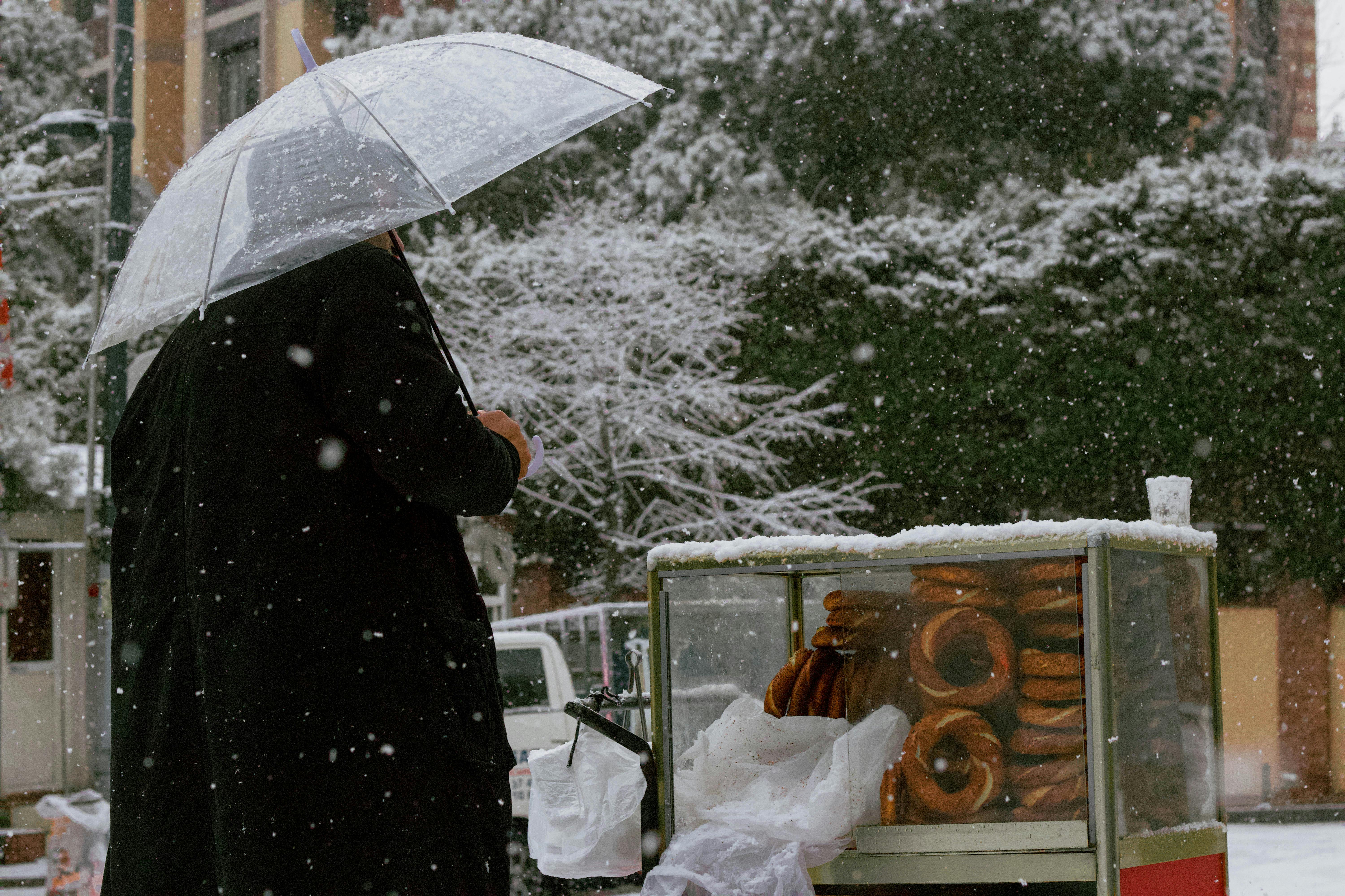 Man with umbrella at street food cart selling simit in snowy Istanbul.