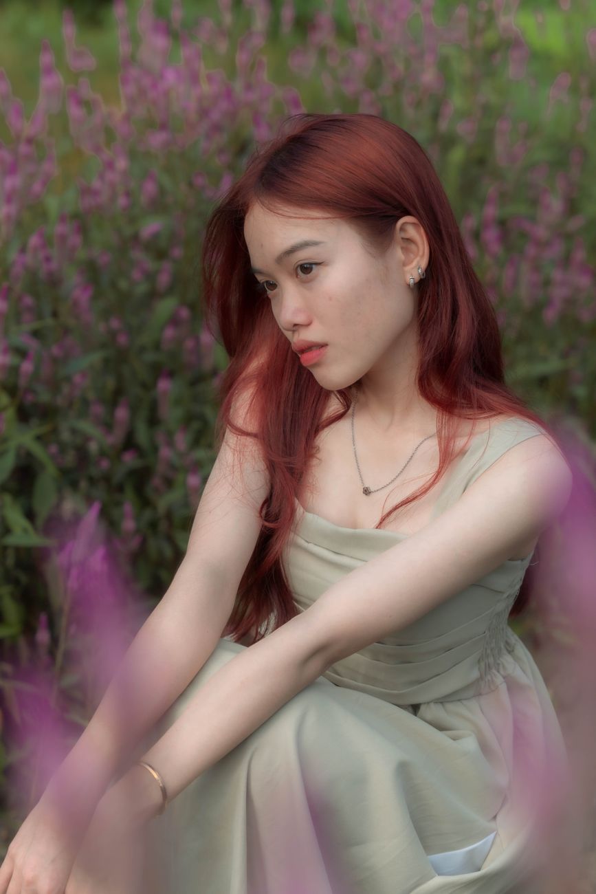 A young woman with flowing red hair sits gracefully among purple flowers.