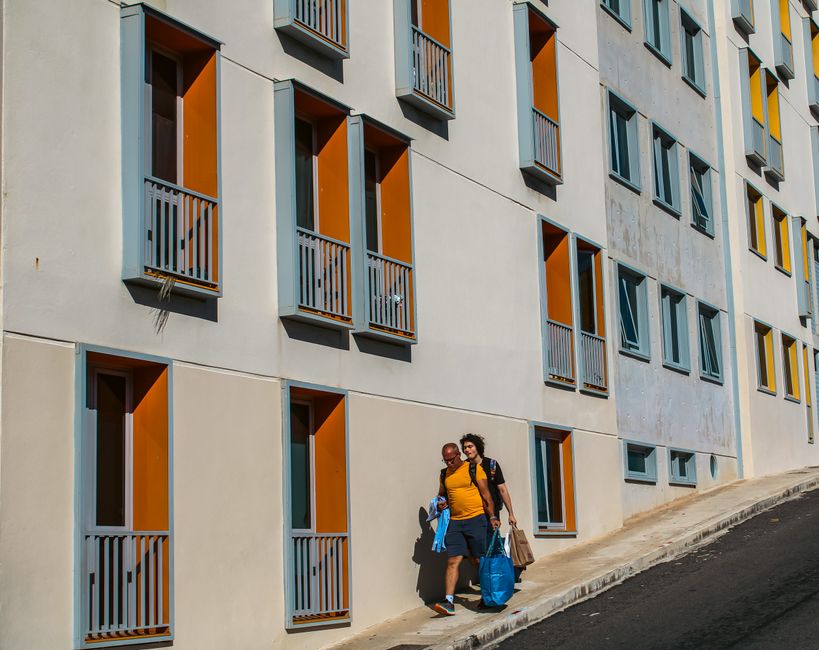 Dos personas caminando junto a un edificio moderno con acentos naranjas en un día soleado.