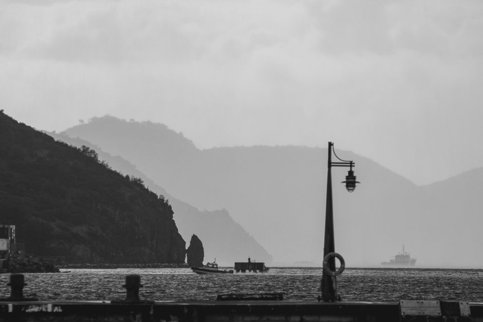 Black and white view of mountains, sea, and a boat, featuring a streetlight silhouette.
