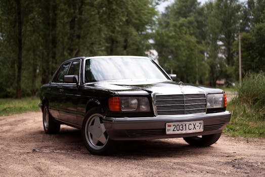 A classic black Mercedes Benz parked on a scenic dirt road in a lush green forest setting.