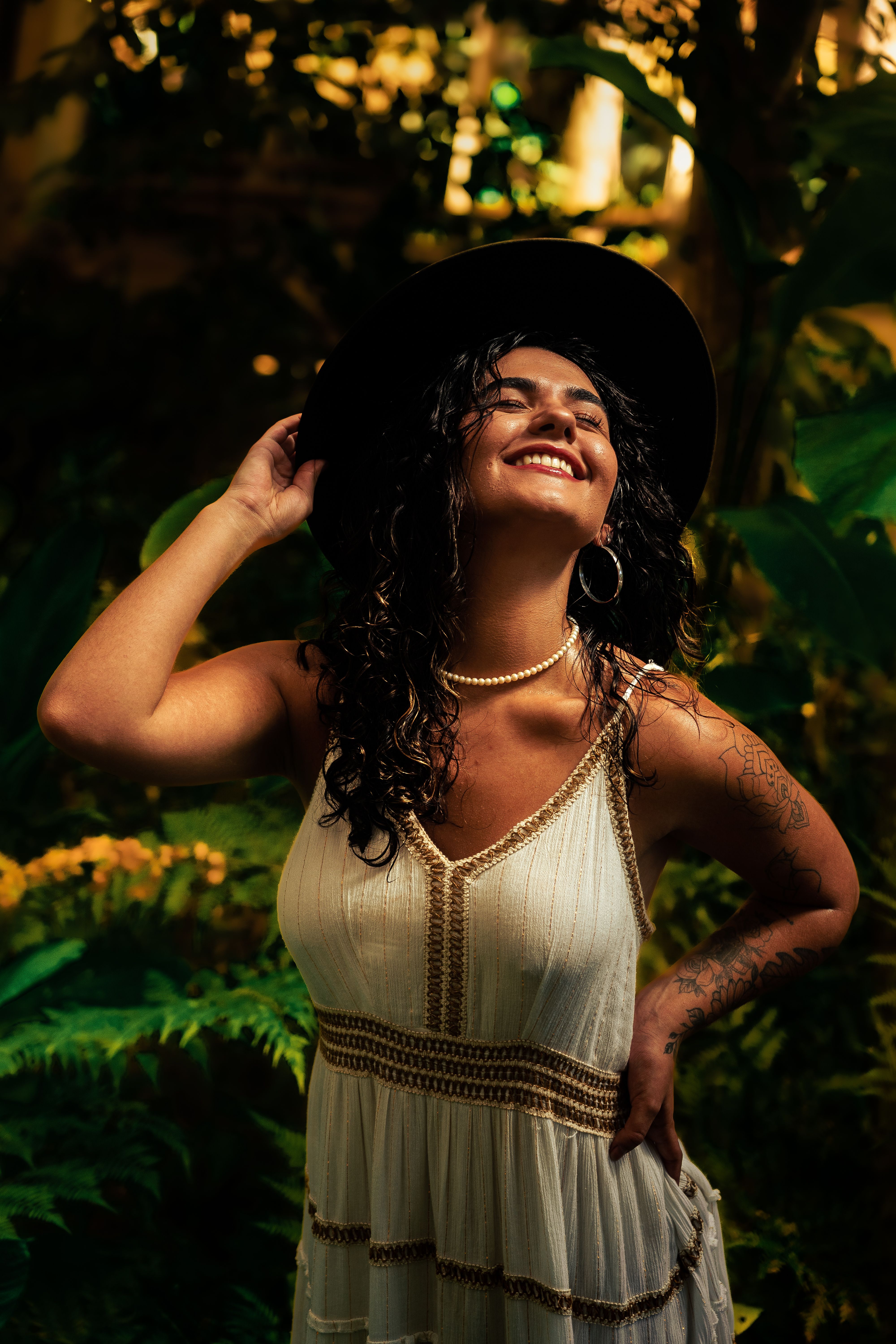 Radiant woman smiling in a white dress and hat surrounded by lush greenery.