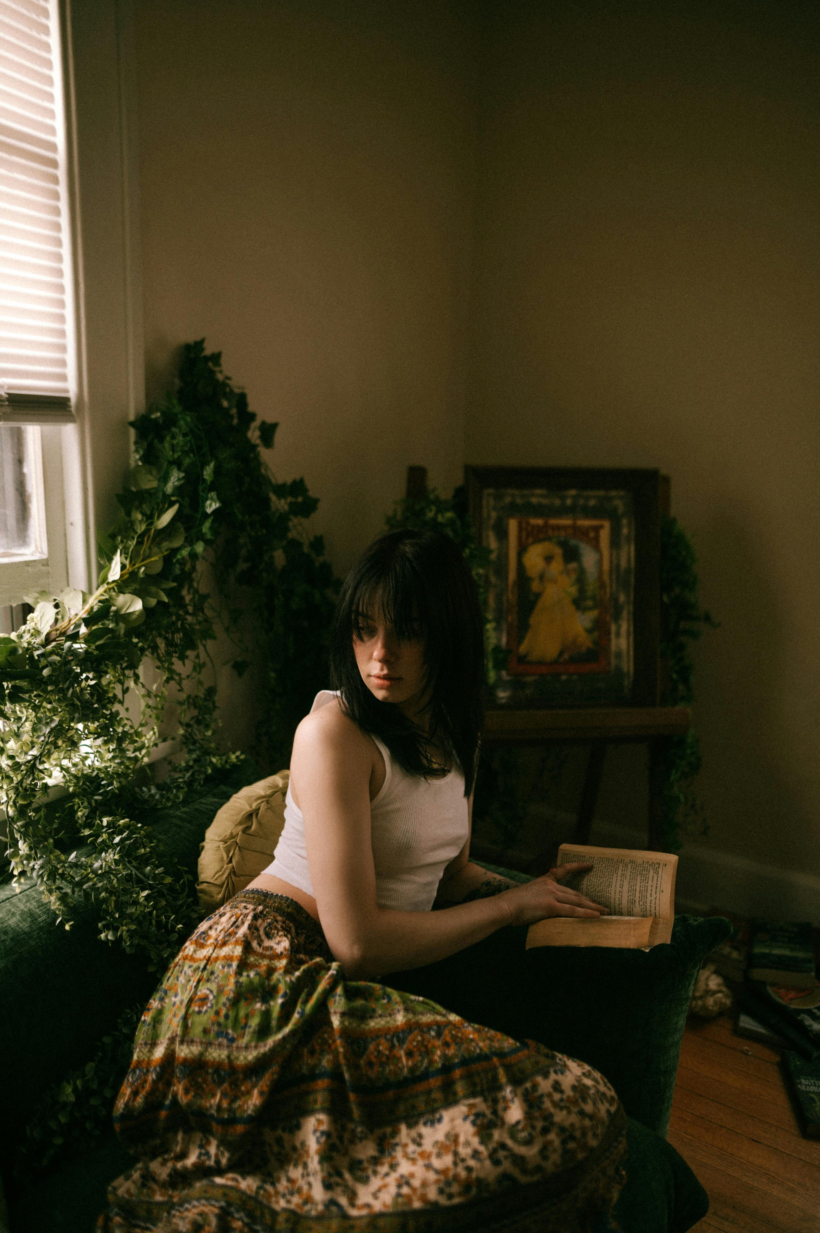 A woman in a patterned skirt reading a book in a cozy, plant-filled room.