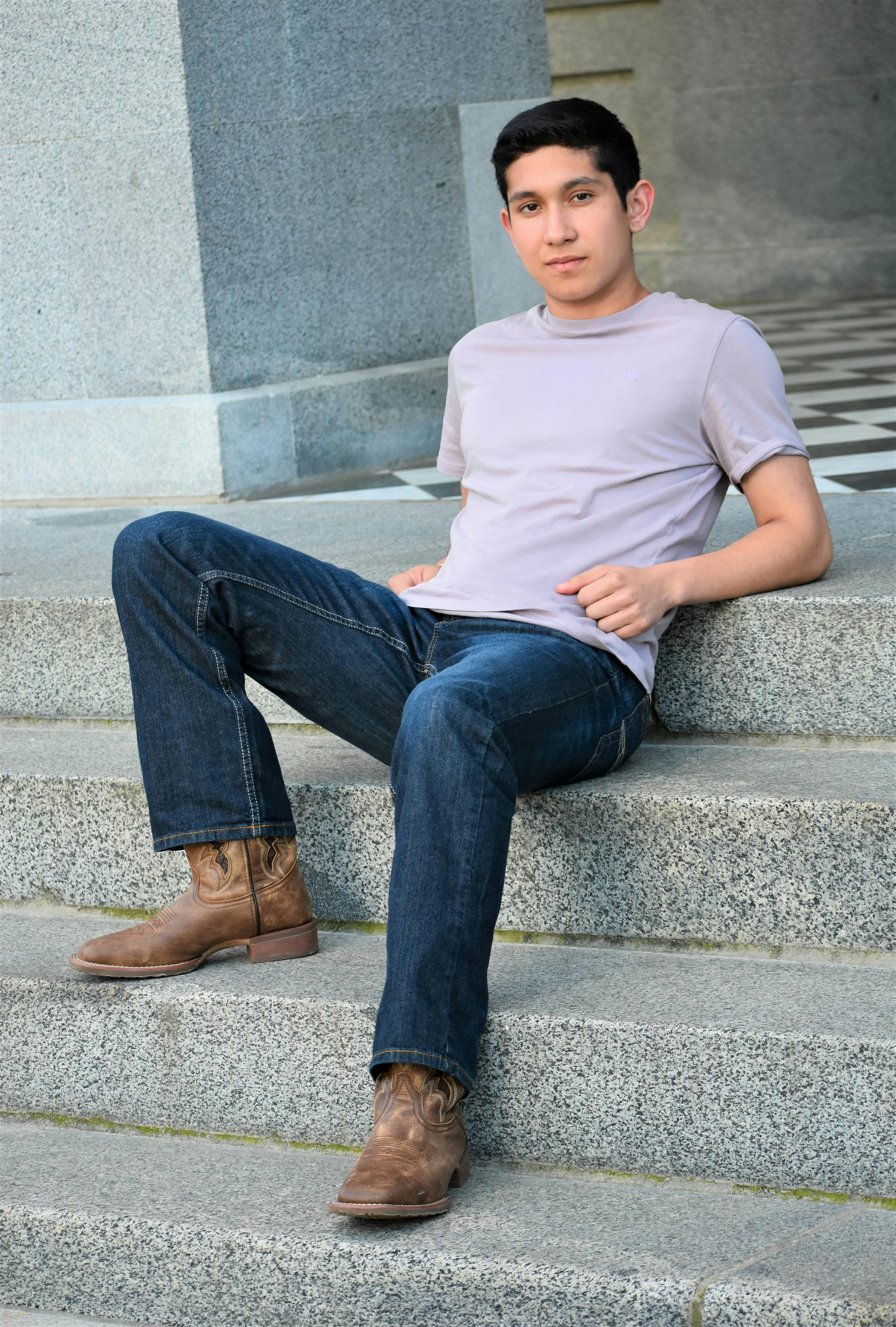 Young man in casual attire sits on stone stairs in an urban setting, showcasing a relaxed and stylish look.
