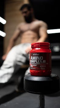 Red amino supplement bottle on dumbbell, blurred muscular man in background.