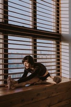 Woman practicing yoga stretch on a wooden windowsill with sunlight streaming in.
