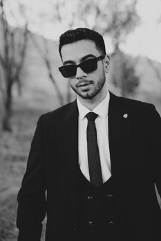 Black and white portrait of a stylish man in a suit and sunglasses outdoors.