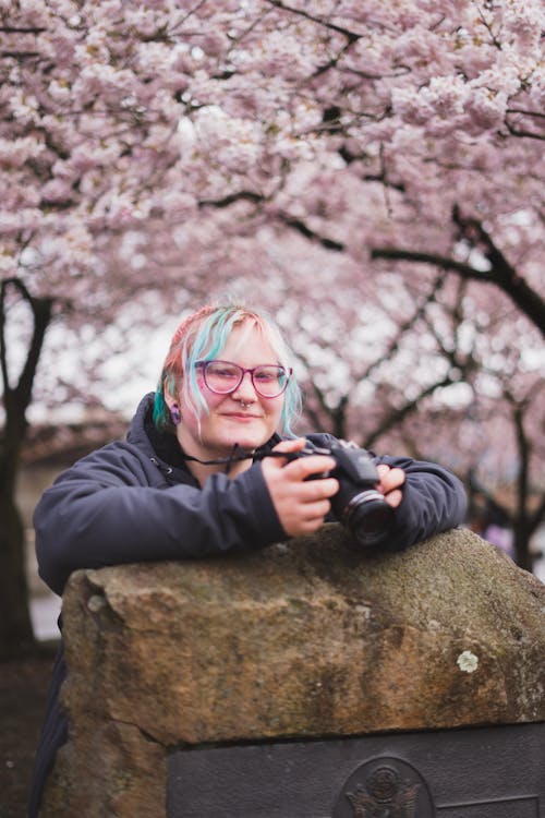 Smiling Woman with Camera Posing with Cherry Trees behind
