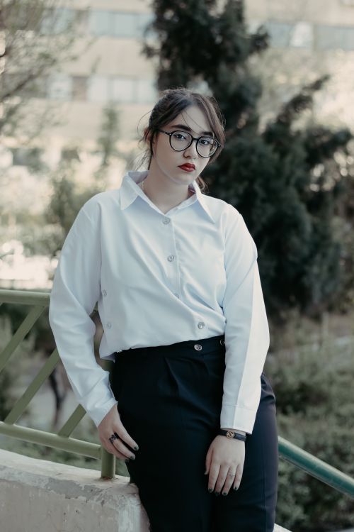 Free Portrait of a fashionable woman in eyeglasses, posing outdoors in a white shirt and black pants. Stock Photo