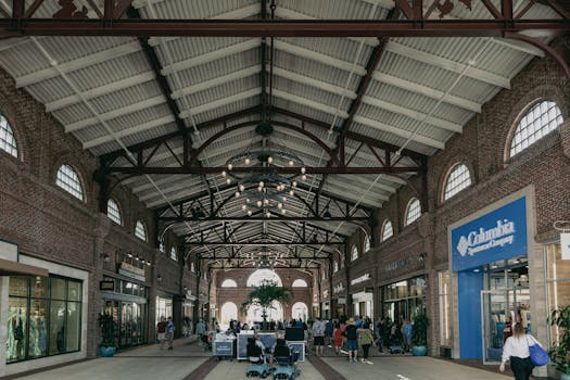 Urban shopping mall interior with people walking amid stores under decorative lighting and exposed beams.