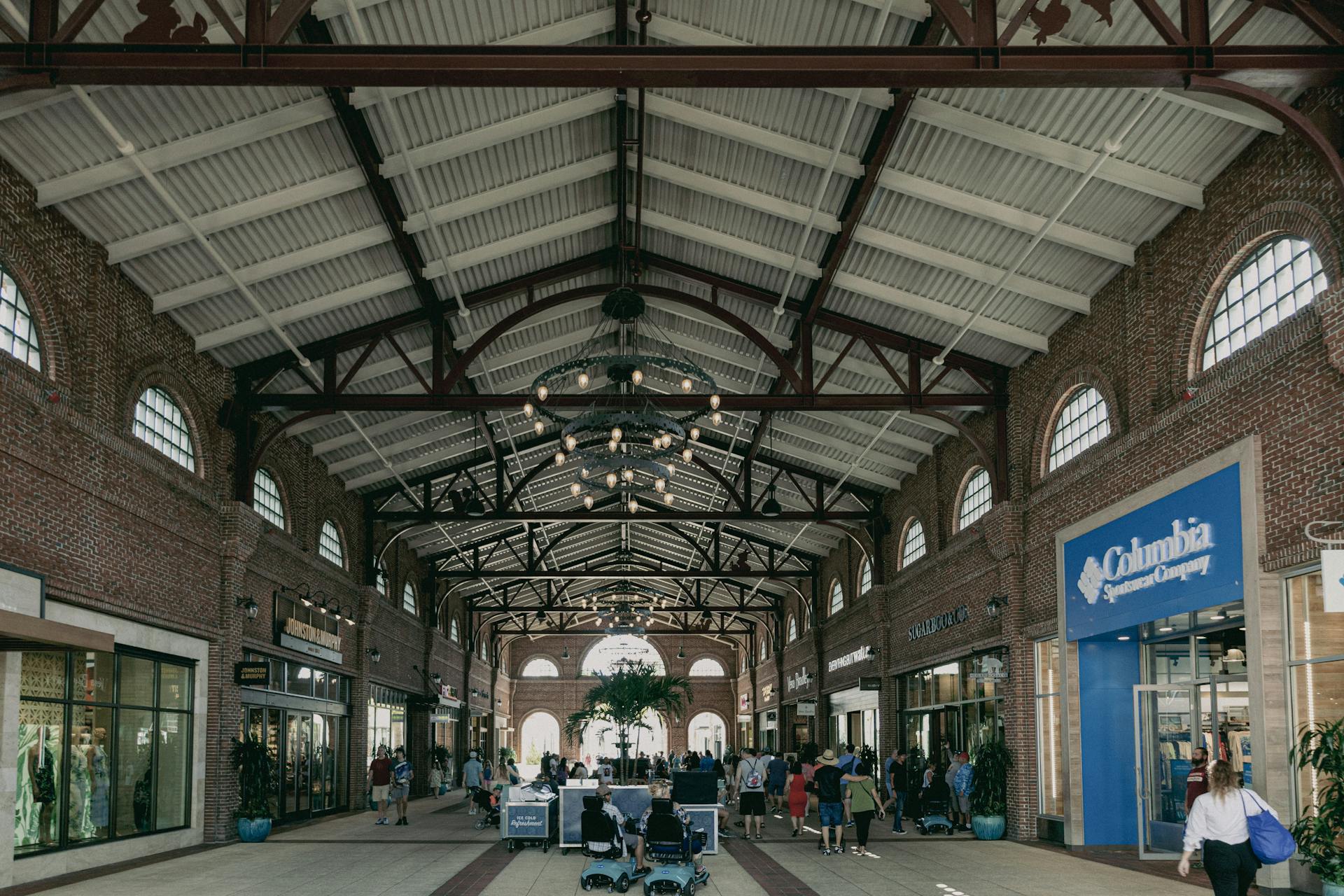 Urban shopping mall interior with people walking amid stores under decorative lighting and exposed beams.