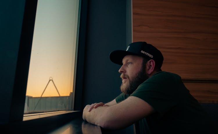 Man In Cap And With Beard Leaning On Windowsill