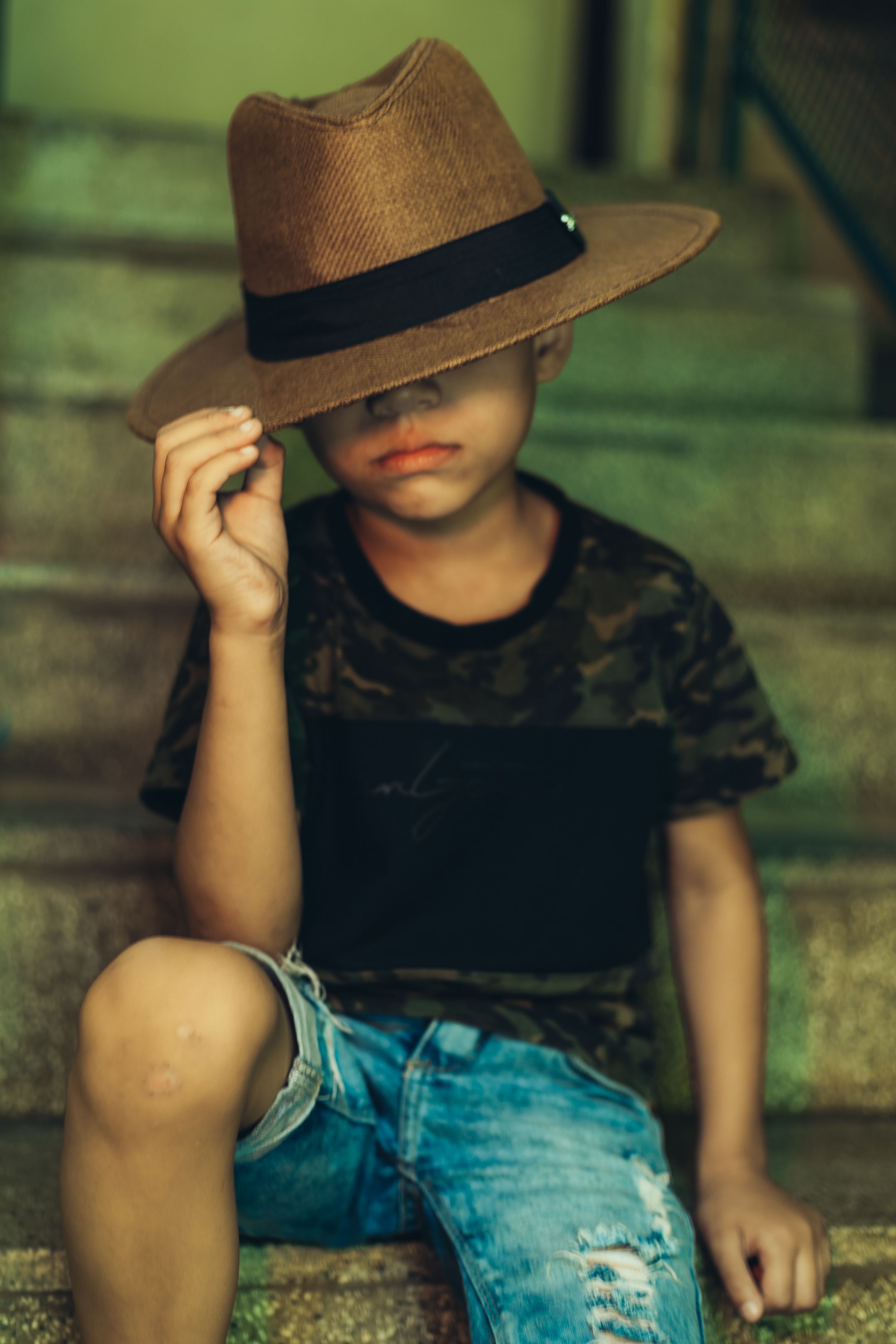 Criança com camisa de camuflagem e jeans posa com chapéu em escadas para retrato estiloso.
