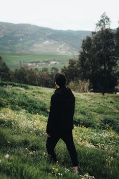 A man in a black coat stands in a lush green field in Constantine, Algeria, admiring the scenic beauty.