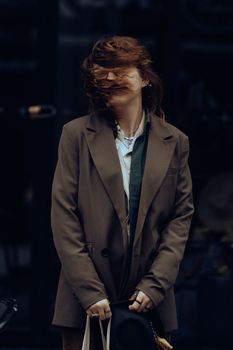 A fashionable woman stands outdoors with tousled hair blowing in the wind, wearing a brown jacket.