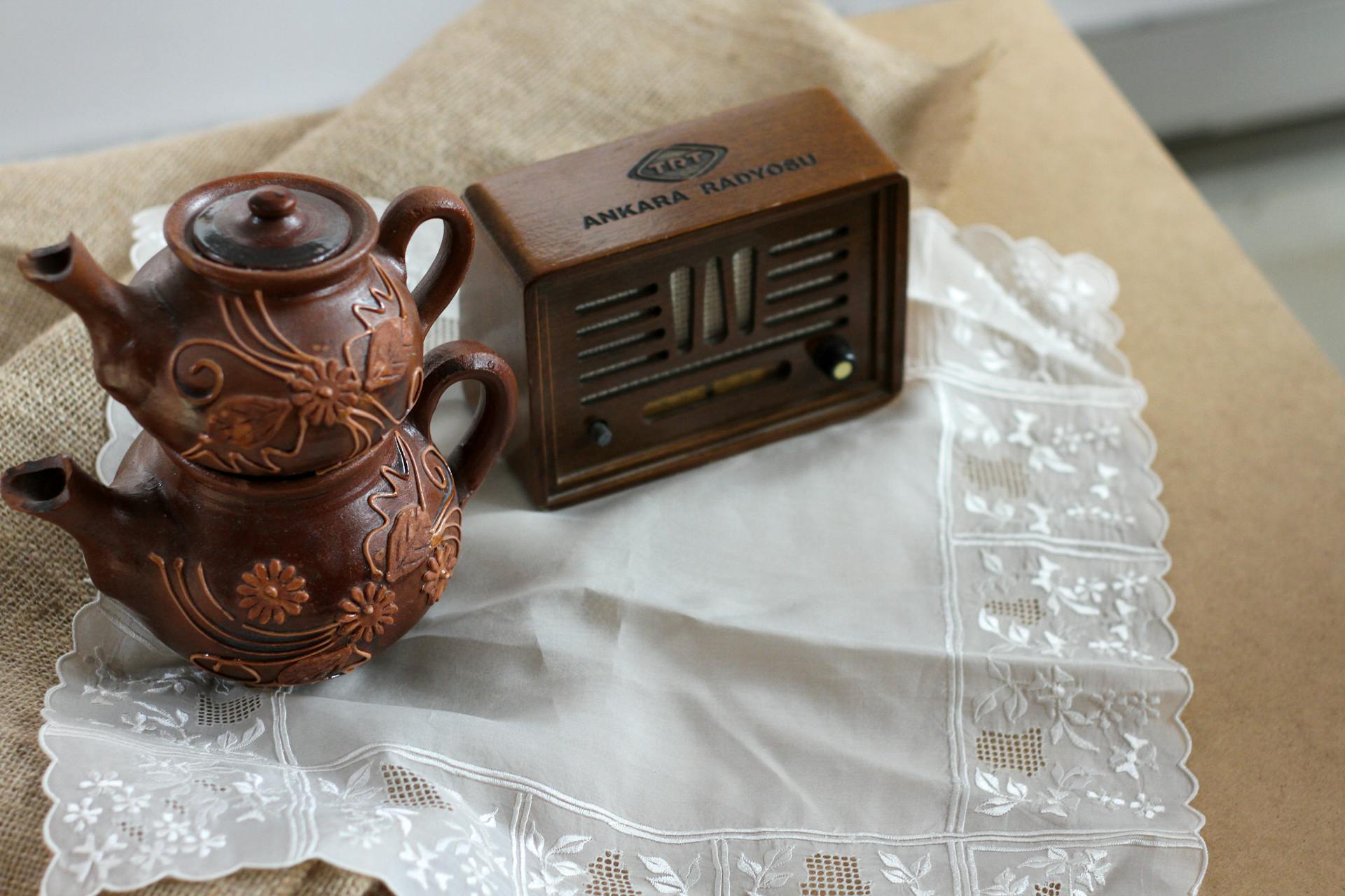 Two stackable vintage teapots with floral design alongside an antique radio on lace cloth.