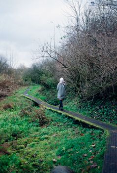 A woman walks on a narrow path surrounded by lush greenery in a serene forest setting.