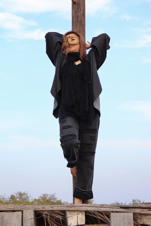 Woman in Mask and Black Clothes Posing by Wooden Pole