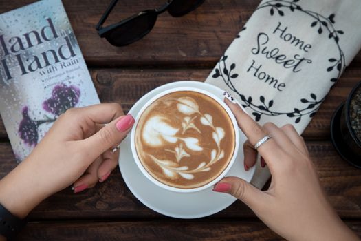 A beautifully crafted latte with floral froth art, accompanied by a book and sunglasses on a rustic table.
