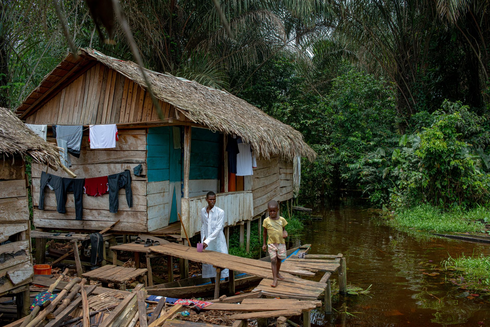 A wooden cabin next to a stream in a lush Nigerian forest with two people present.