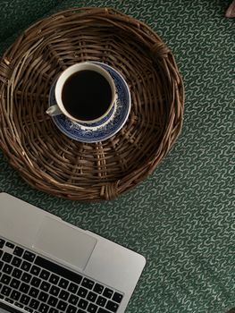 Top view of a cup of coffee on a wicker tray beside a laptop, creating a cozy workspace vibe.