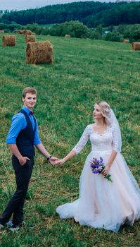Newlyweds holding hands in field with hay bales, embracing love in nature.