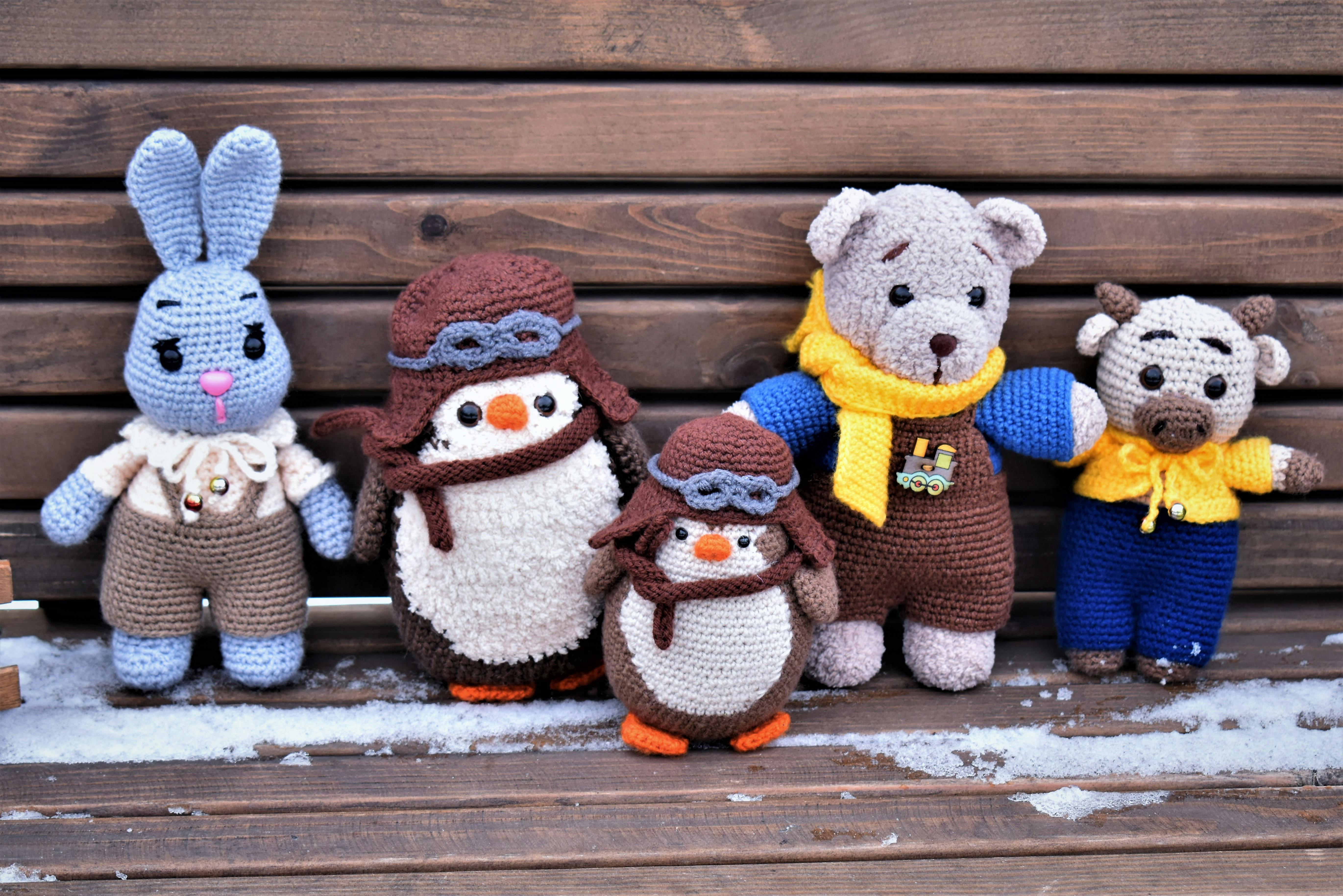 a few winer related crocheted animals sitting on a snowy bench, the animals are a rabbit, two penguins, a bear, and a cow, in winter attire.