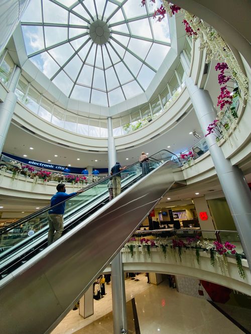 Free A busy, modern shopping mall with escalators and glass dome ceiling, vibrant floral decor. Stock Photo