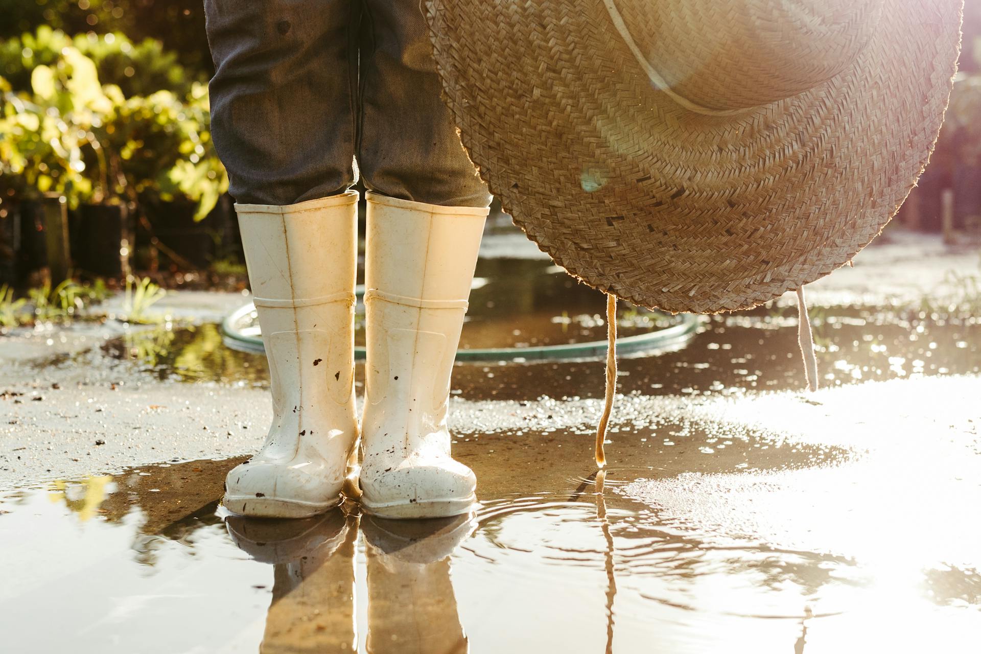 Close-up of a farmer's white boots and straw hat reflecting in a sunny puddle outdoors.