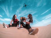 在明亮的蓝天下，两名妇女在迪拜的沙丘上控制着一架无人机