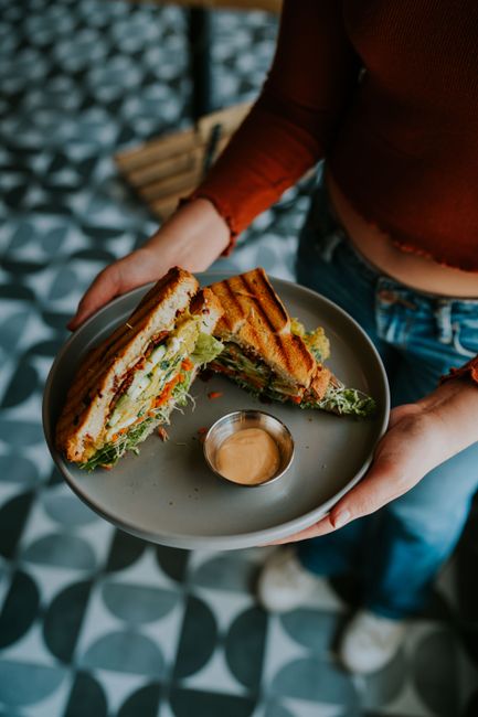 Close-up of a grilled sandwich with lettuce and sauce held by a woman over patterned floor.