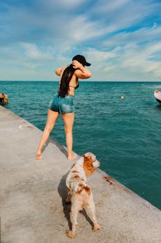A woman in summer attire stands by the sea with a dog on a sunny day.
