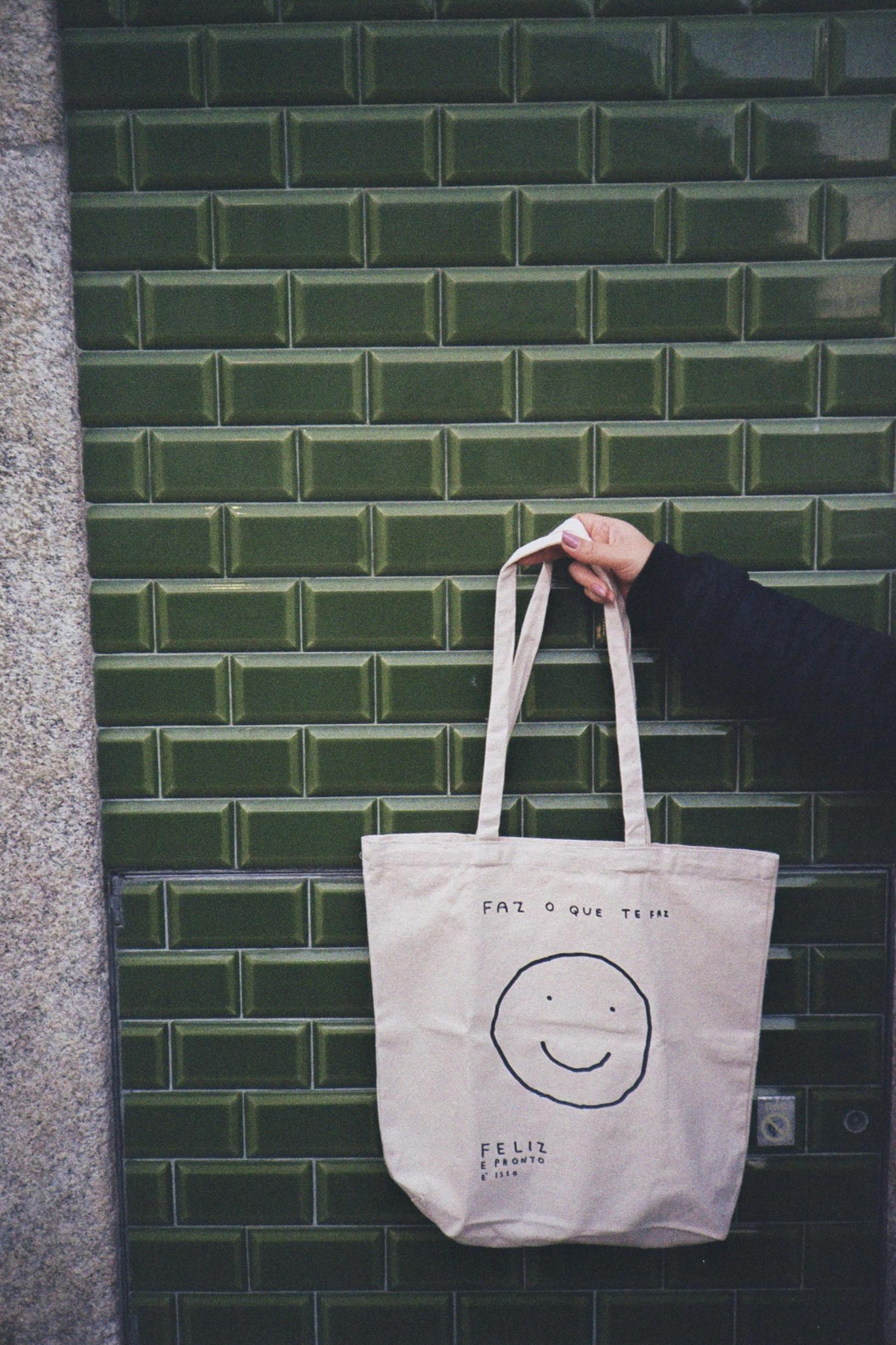 Hand holding a canvas tote bag against a green tile wall. Stylish and eco-friendly accessory.