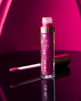 Close-up of a pink glittery lip gloss with a luxury feel, featuring a glass bottle on a vibrant pink background.