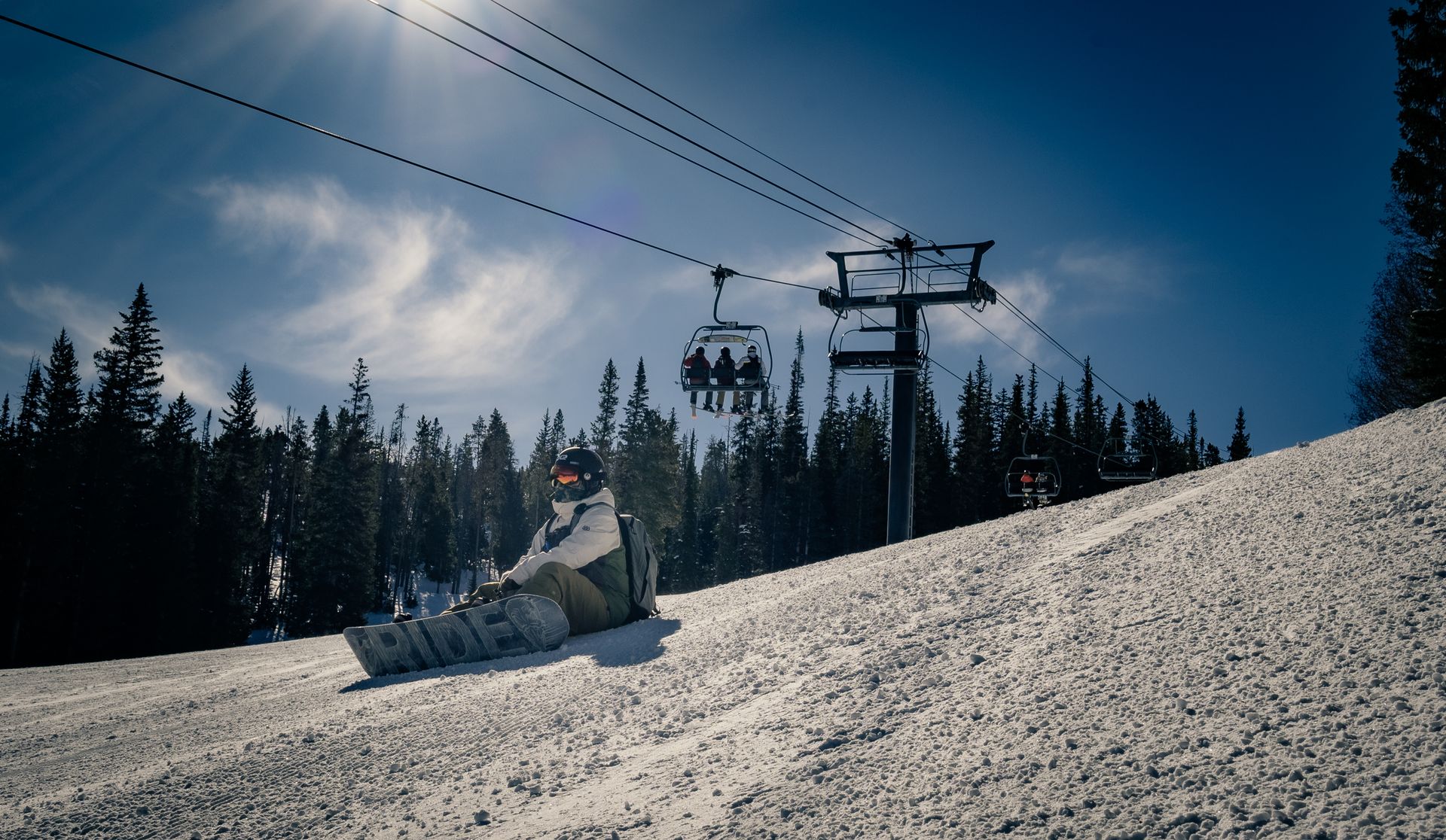 Snowboarder sits on snowy slope under bright sun, ski lift in background.