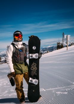 Snowboarder standing on a groomed ski slope holding a snowboard in bright winter gear.