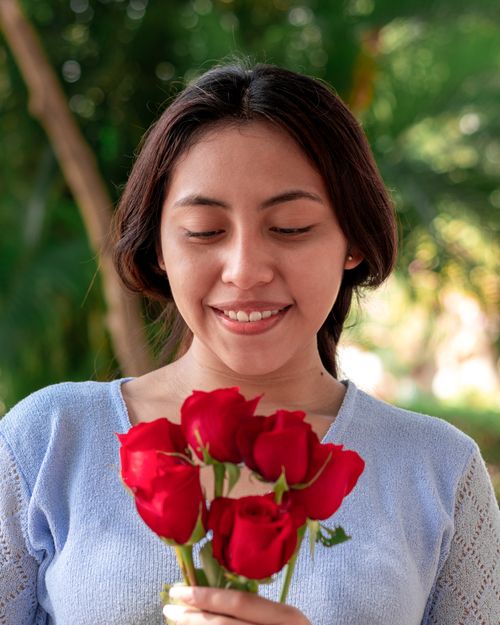 Free A young woman smiles while holding vibrant red roses in an outdoor garden setting. Stock Photo