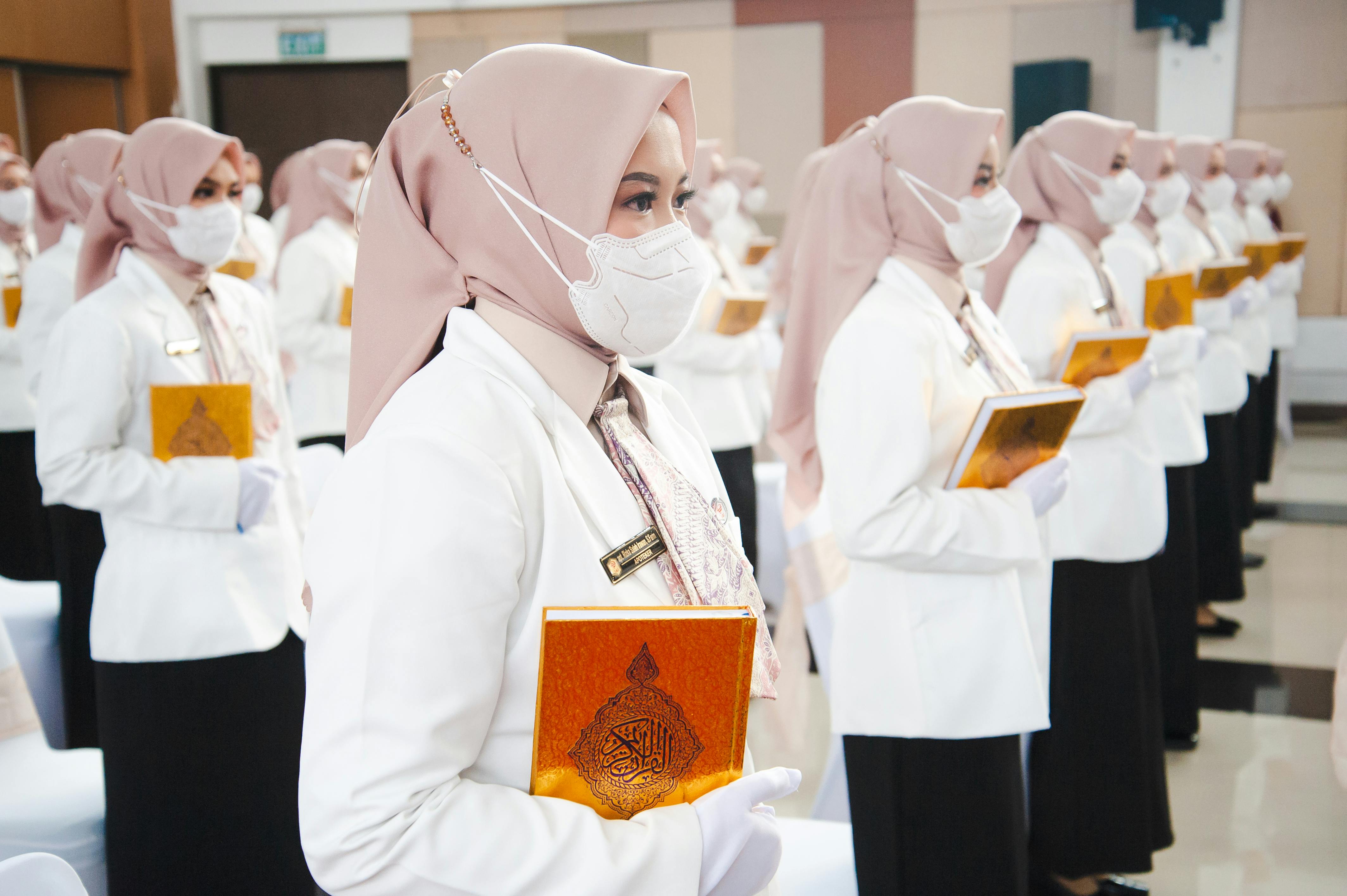 Muslim women in hijabs holding certificates during a graduation ceremony, all wearing masks and formal attire.