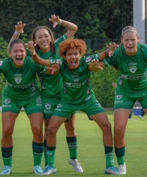Four young female soccer players in green jerseys celebrate energetically on a sunny outdoor field.