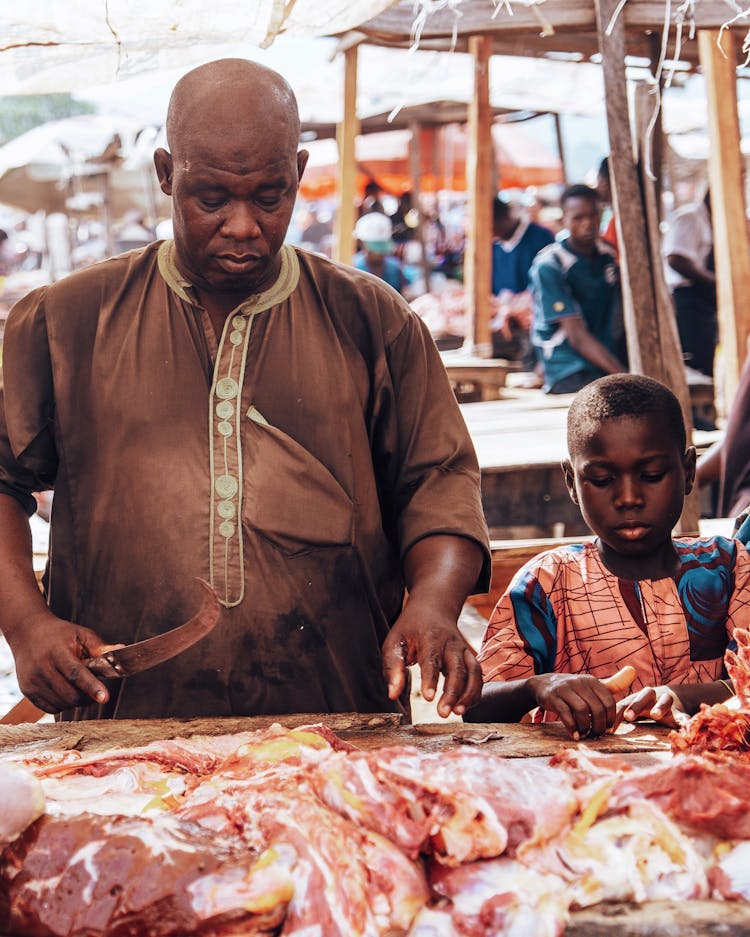 Father And Son Standing By Butcher Stand At Bazaar