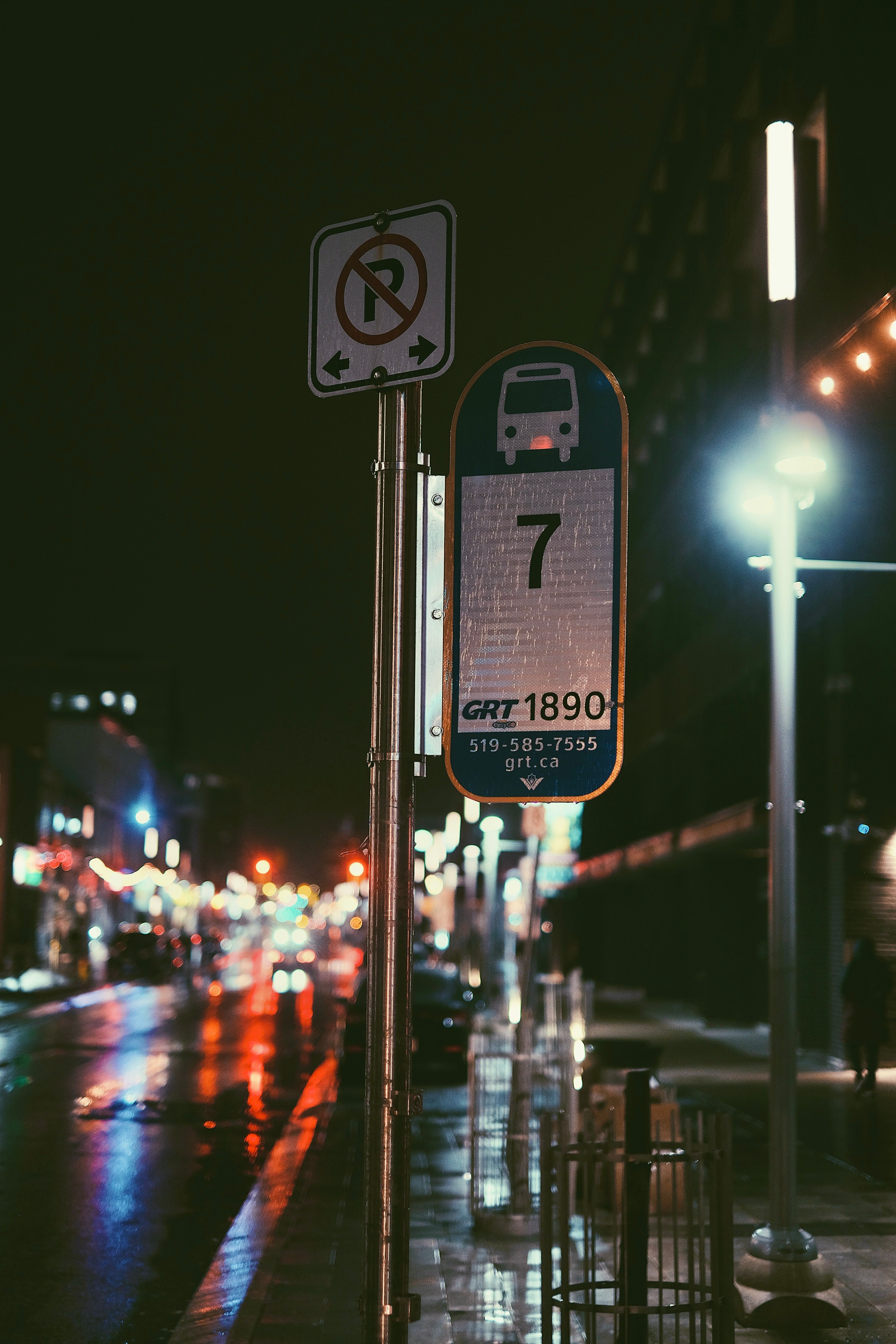 Moody night scene of a Kitchener bus stop reflecting city lights on wet streets.