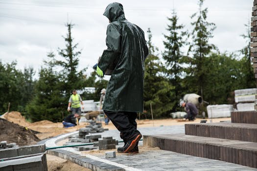 Worker in raincoat laying pavement at outdoor construction site on a rainy day.