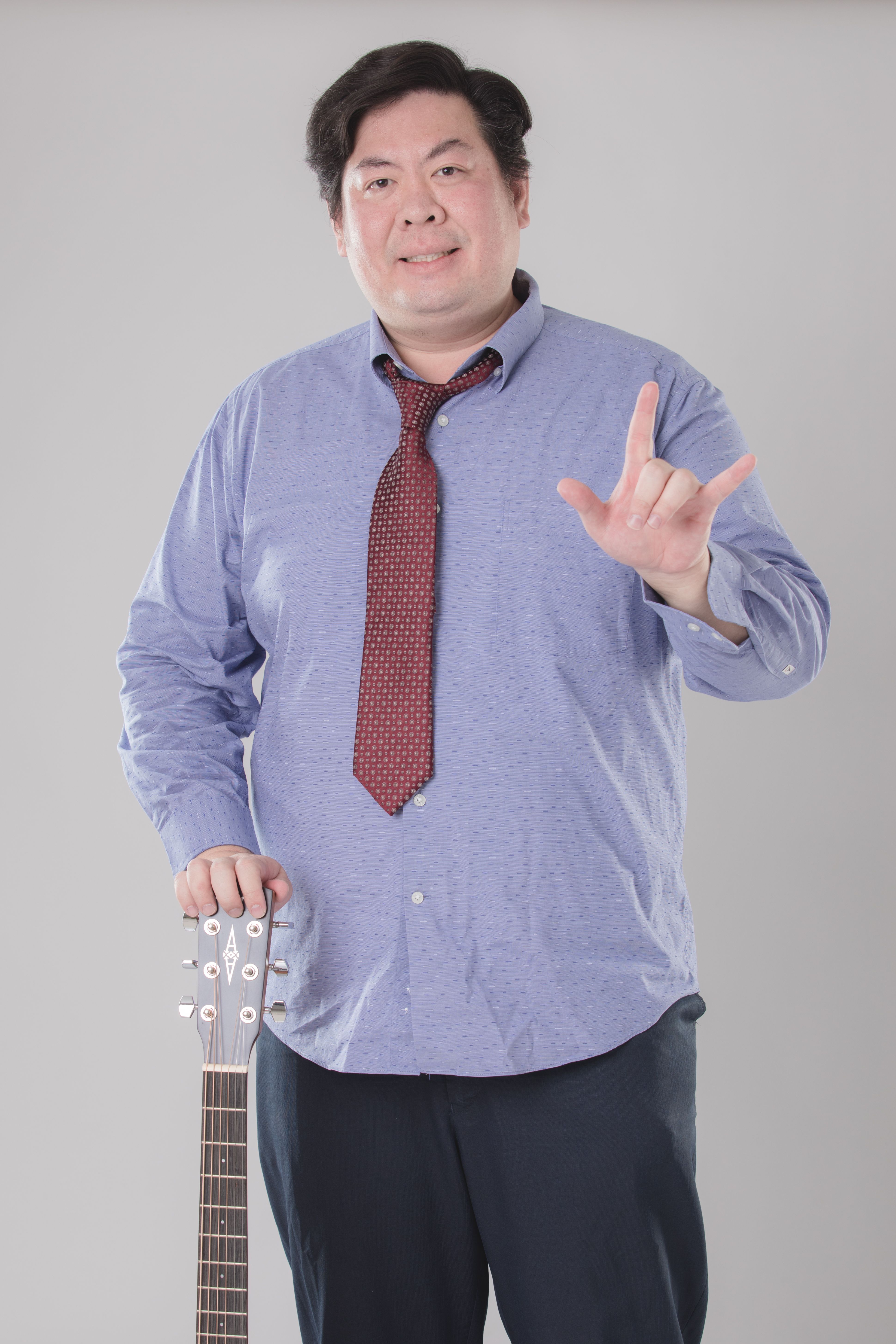 Casual portrait of an adult musician holding a guitar, wearing a blue shirt and red necktie.