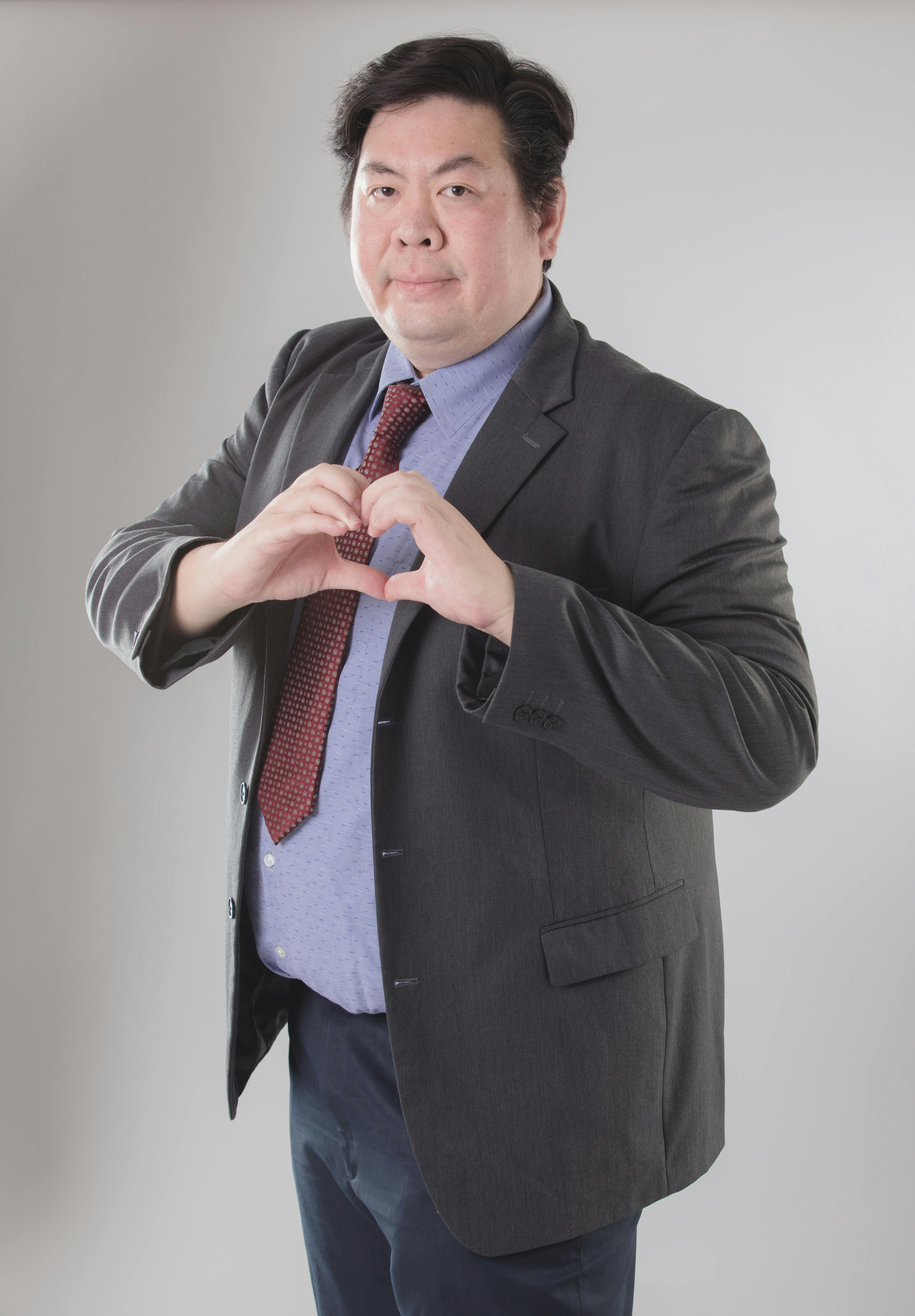 Adult male in a suit and tie making a heart gesture with hands in a studio setting.