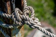 Detailed view of a rope knot tied around a rusted post outdoors under sunlight.