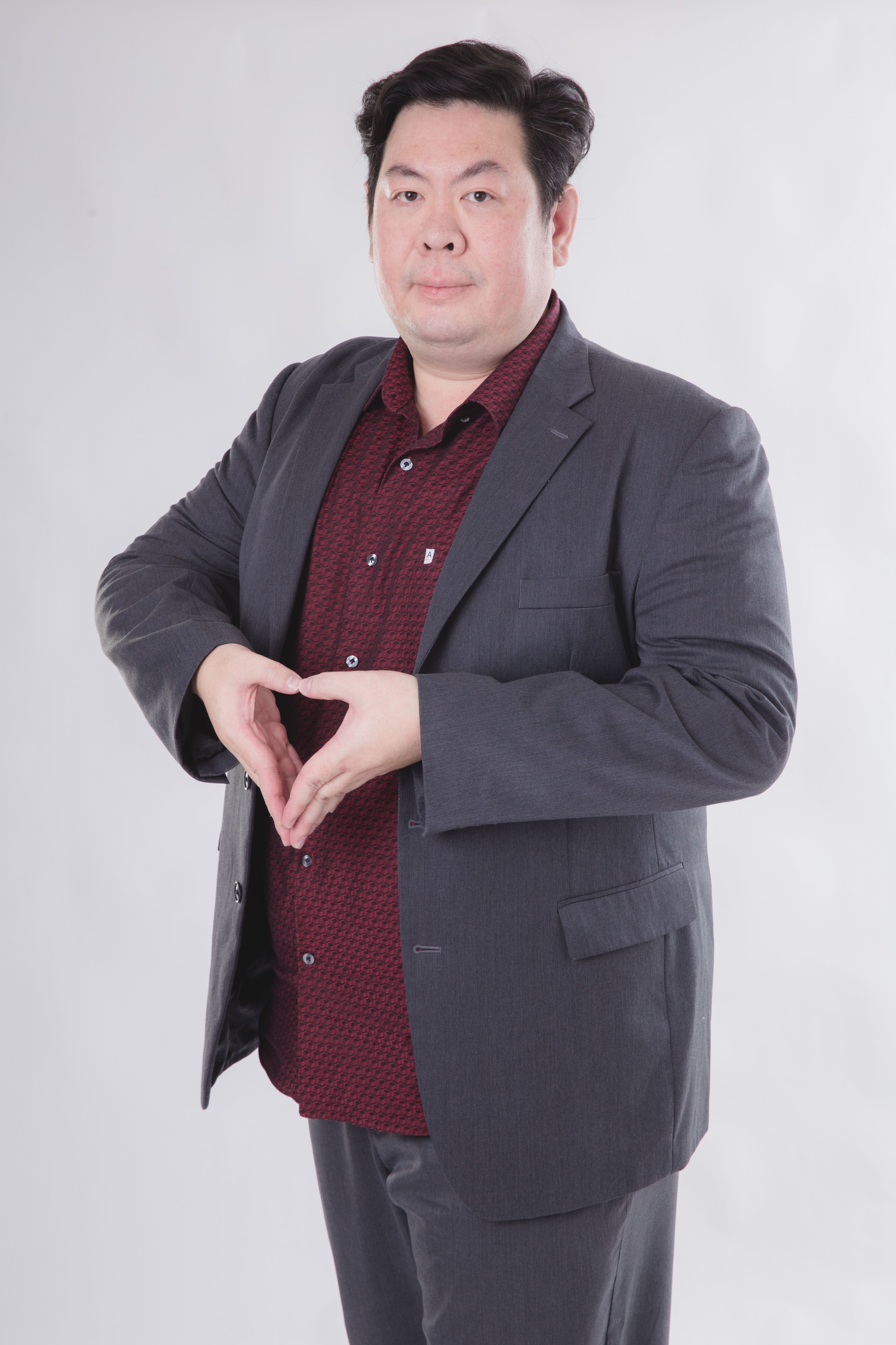 Portrait of an Asian man in a suit making a heart gesture with his hands, standing against a neutral background.