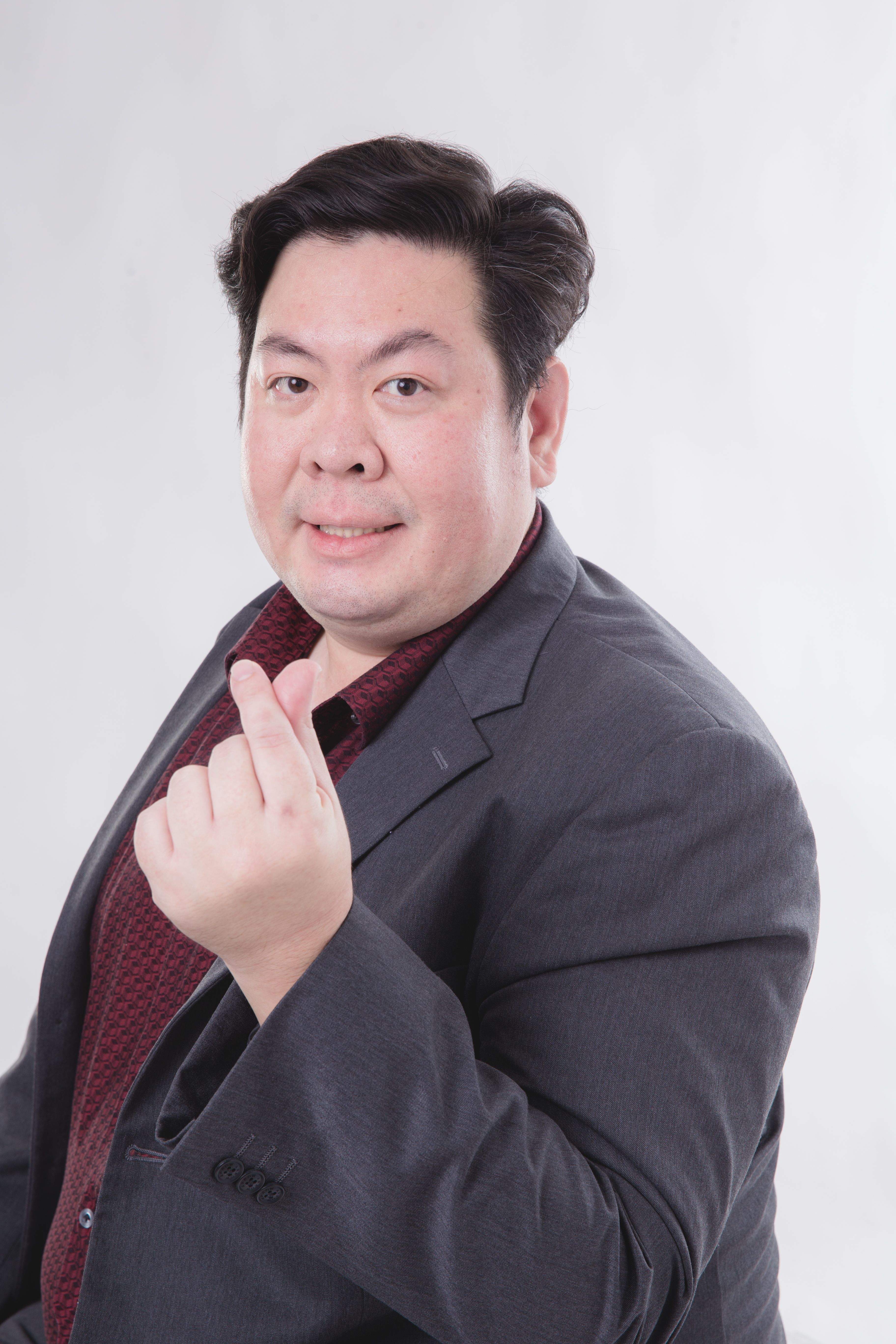 Studio portrait of an adult man in a suit making a heart gesture with his hand, smiling confidently.
