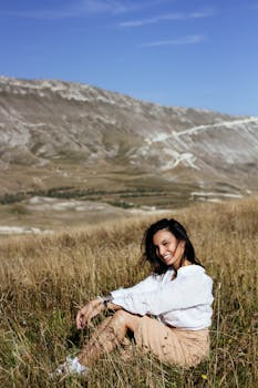 A cheerful woman sits in a sunlit meadow with mountains in the background, embodying nature's beauty.