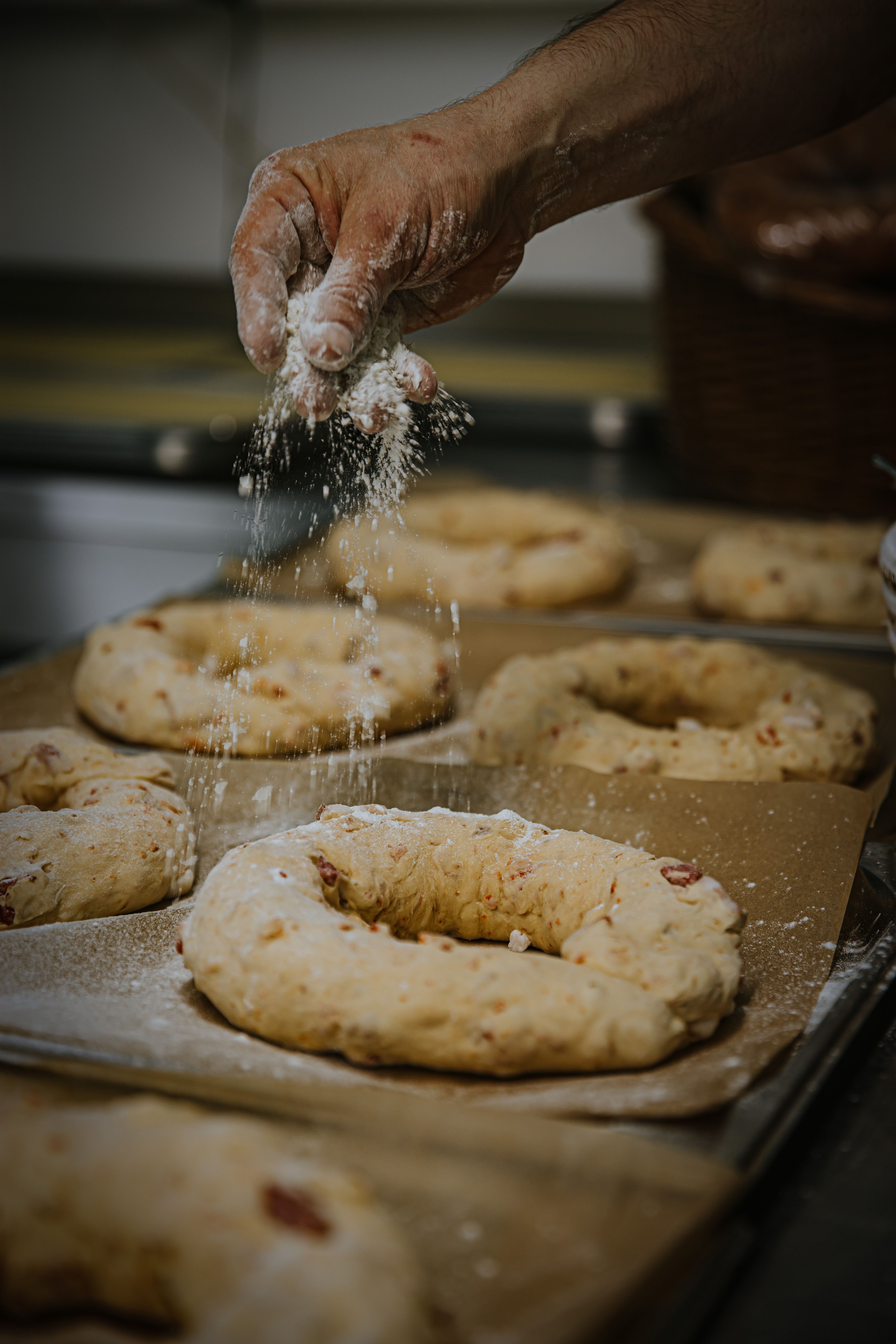 Close-up of a baker's hand sprinkling flour on dough rings in a kitchen setting.